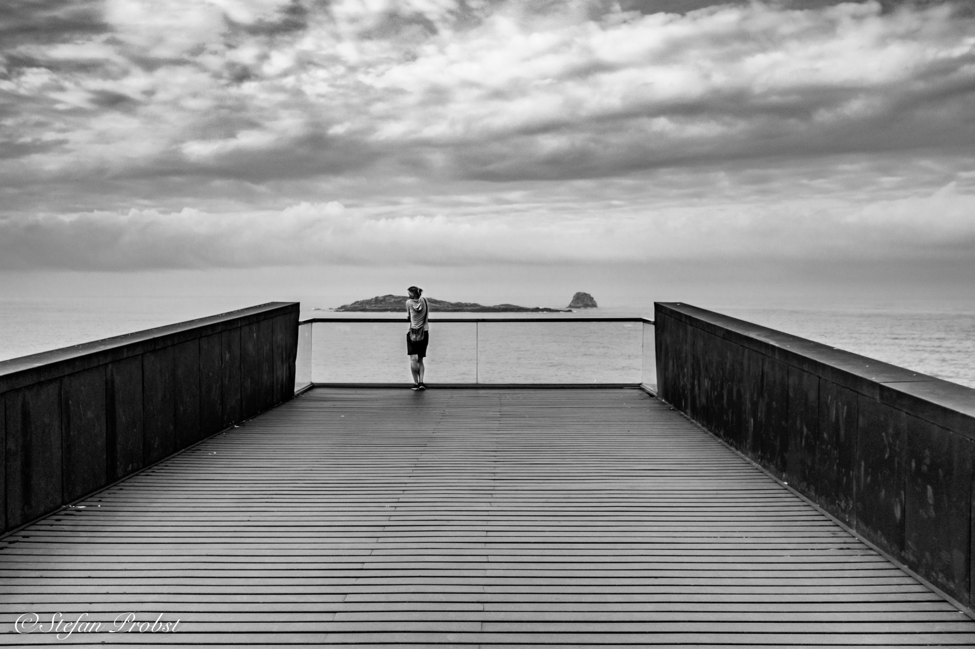 Schwarz-weiß Bild einer Frau auf einem Eisensteg der über dem Meer schwebt.